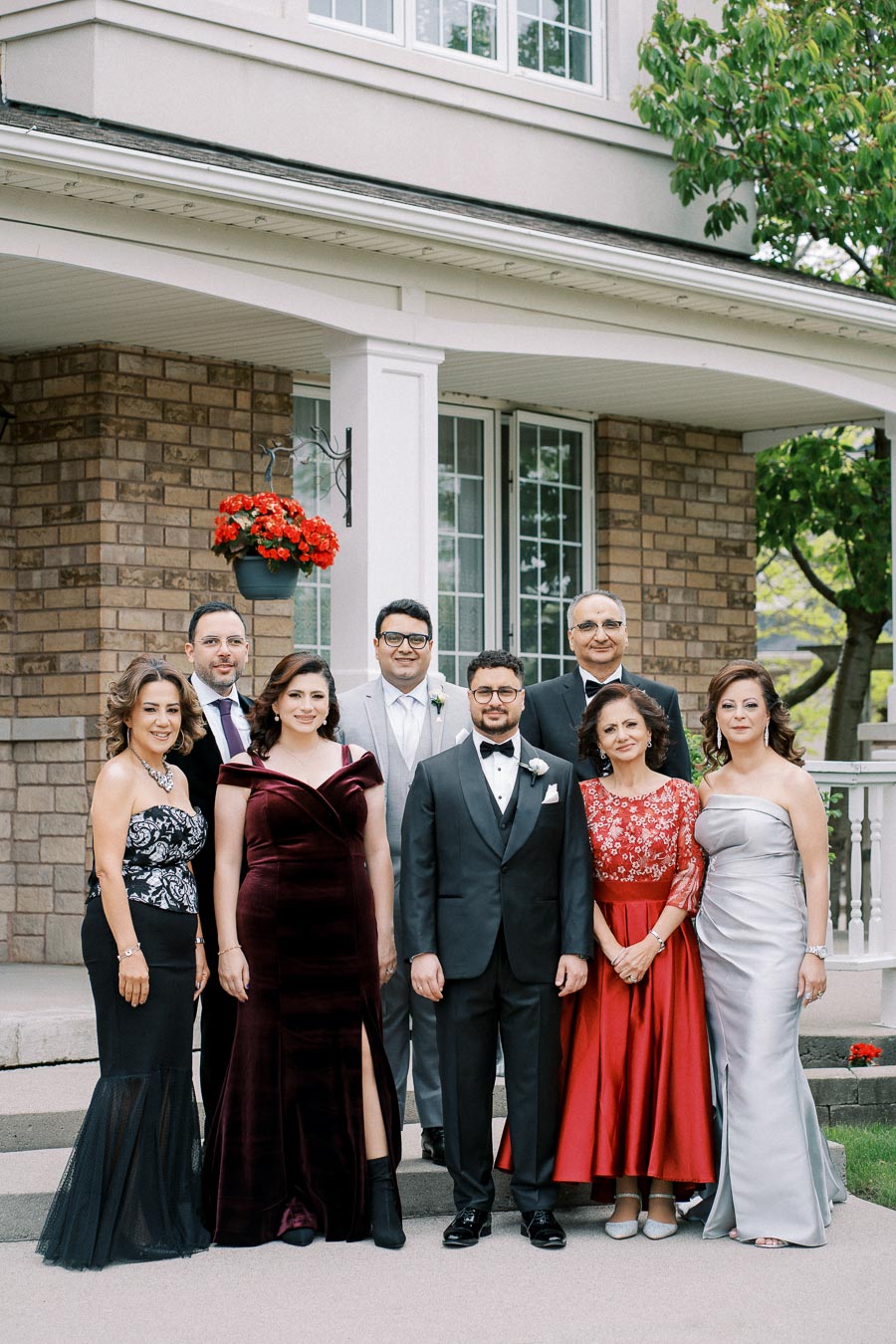 A group of eight well-dressed individuals standing in front of a brick house with a flower pot hanging on the porch.