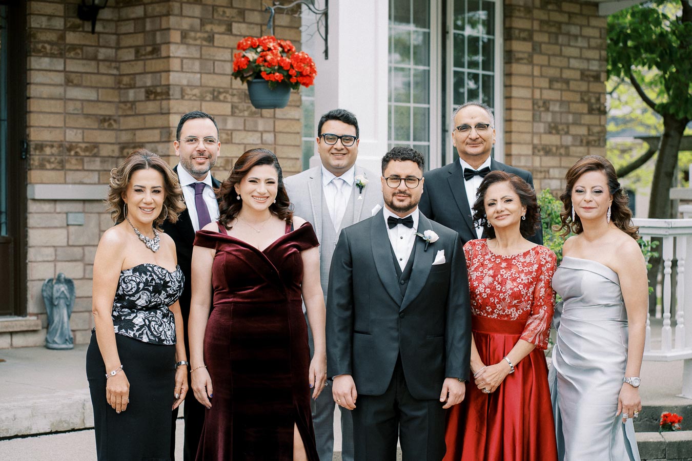 A group of elegantly dressed people posing outdoors, with women in formal dresses and men in suits, standing in front of a brick building with a hanging flower pot.