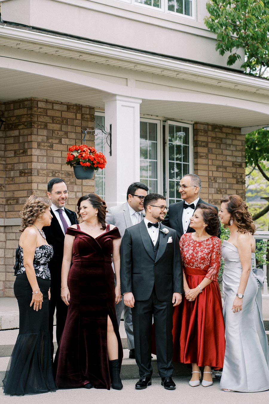 Group of elegantly dressed people posing outdoors at a formal event, standing in front of a brick house with hanging red flowers.