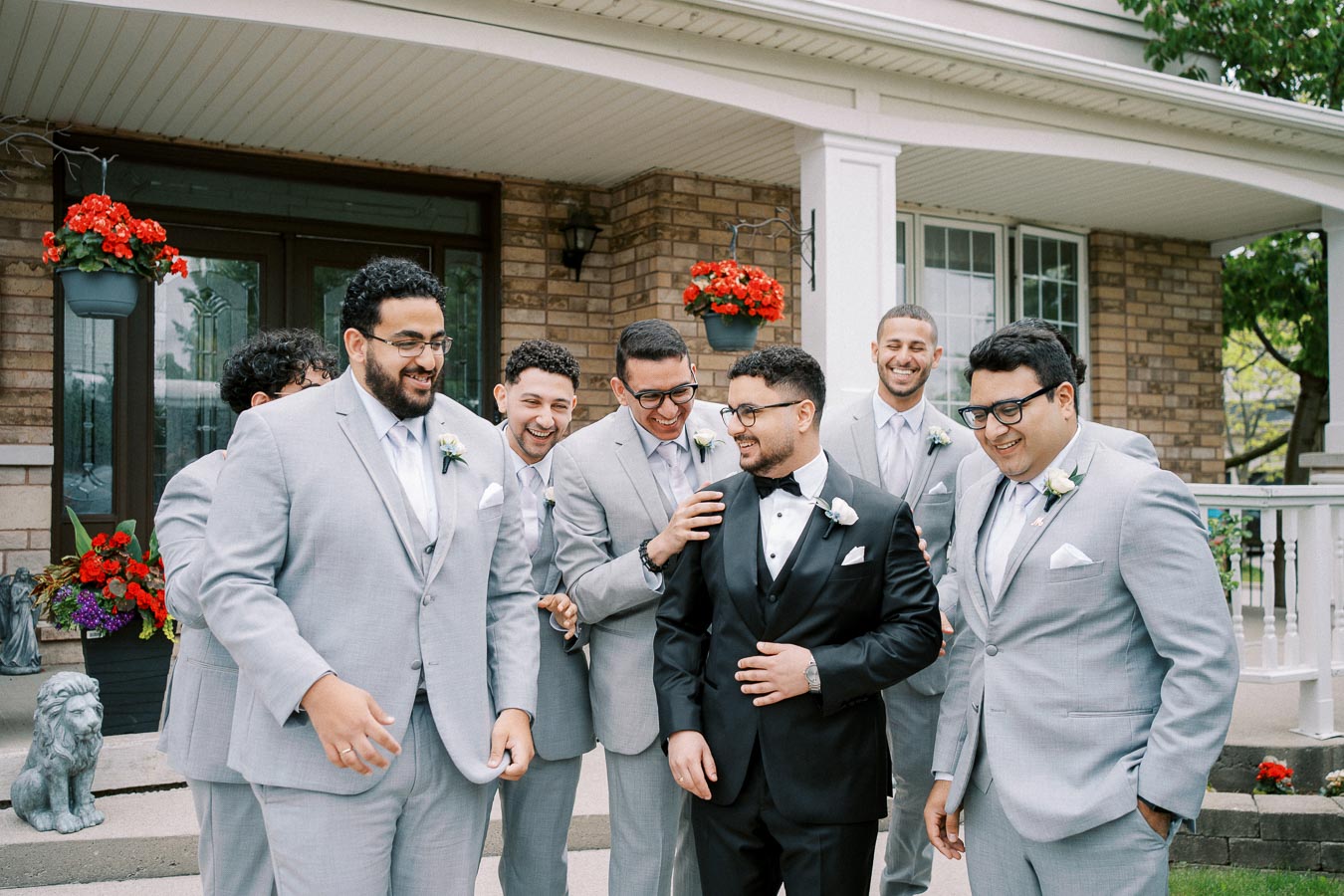 Groom in black tuxedo laughing with groomsmen in light gray suits outside a house, with red flowers in the background.