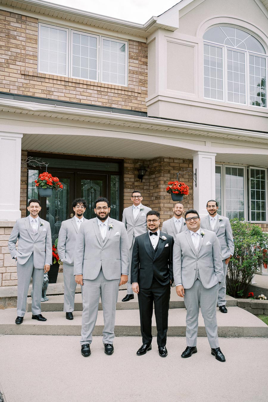 Groom and groomsmen in formal suits posing in front of a brick house with flower pots, showcasing a wedding day celebration.