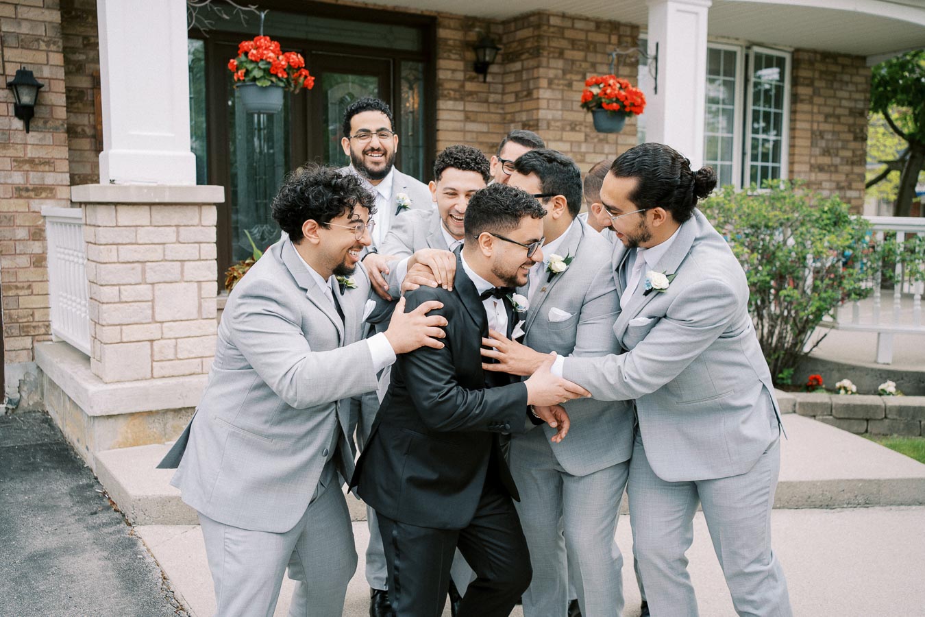 Groomsmen in matching grey suits joyfully surround the groom in black tuxedo on a wedding day, captured outside a brick house adorned with red flowers.