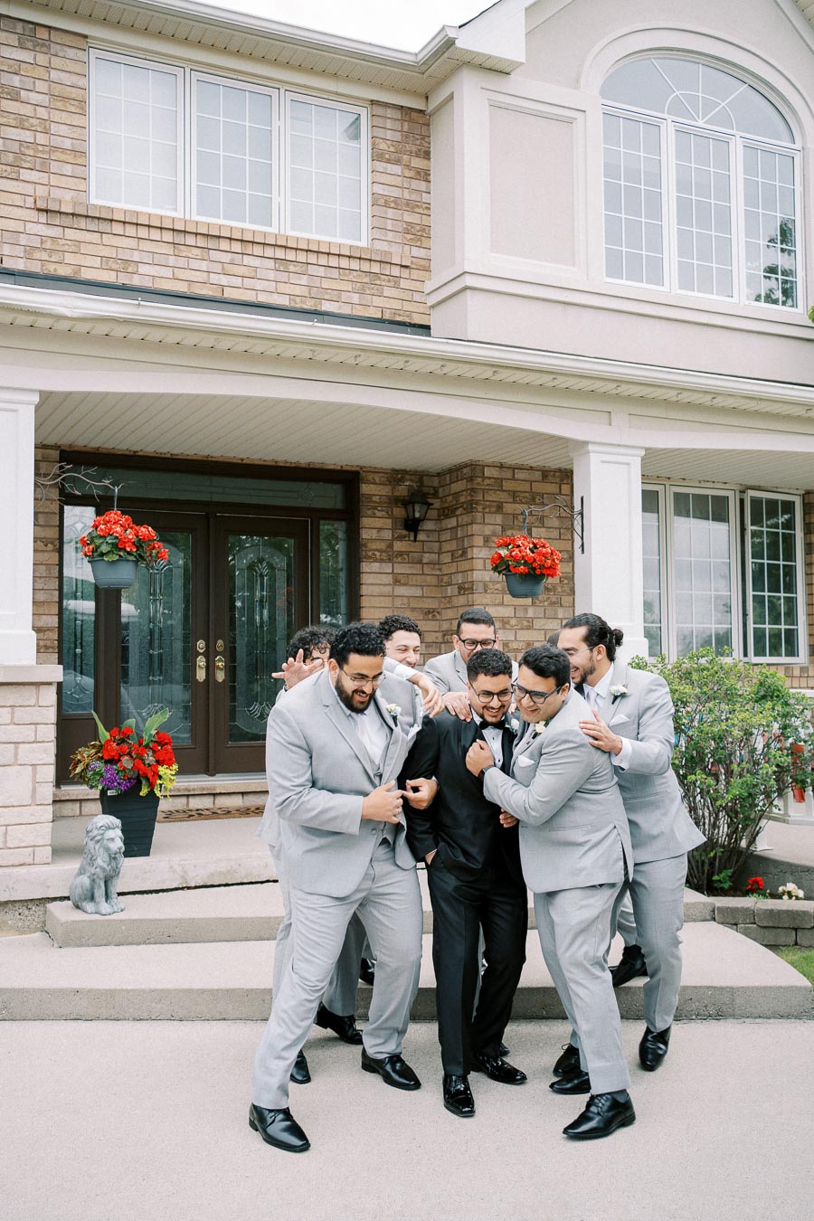 A group of groomsmen in matching gray suits playfully surround the groom, who is dressed in a black tuxedo, in front of a brick suburban house adorned with red flower arrangements.