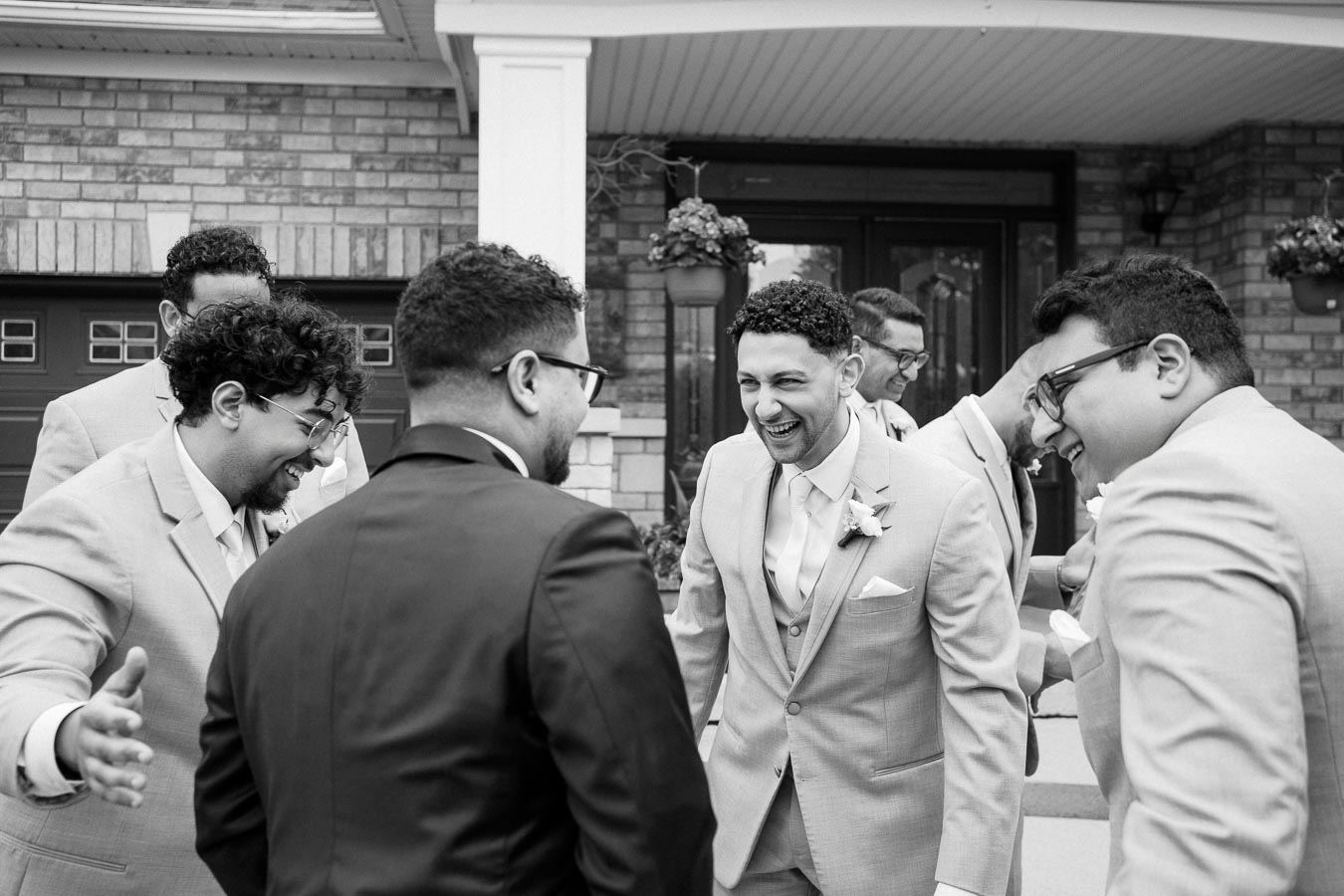A group of joyful groomsmen sharing a laugh in front of a house, dressed in elegant suits, capturing a candid moment before a wedding ceremony.