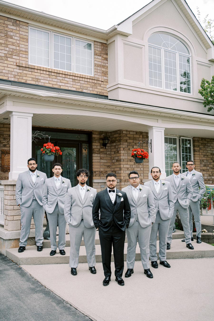 Groom in a black tuxedo standing with groomsmen dressed in light gray suits, posing in front of a brick house with large windows and red flowers.