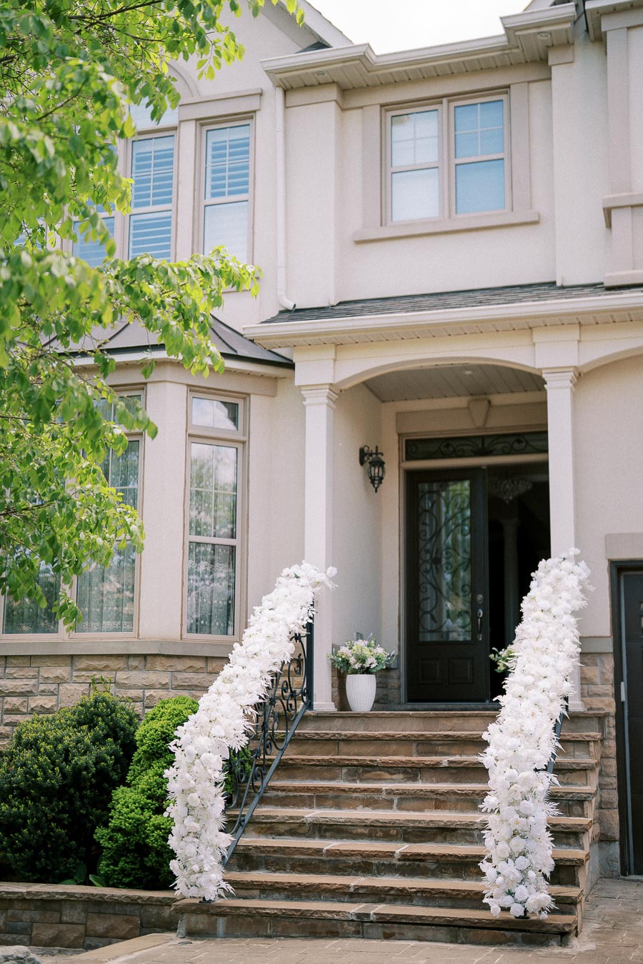 Elegant front entrance of a home decorated with white floral arrangements on the stair railings, surrounded by lush greenery, creating a welcoming and sophisticated atmosphere.