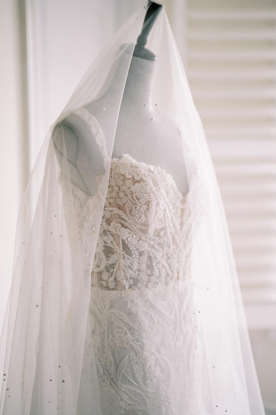 Elegant lace bridal gown with intricate detailing displayed on a mannequin, covered by a delicate tulle veil, in a softly lit room.