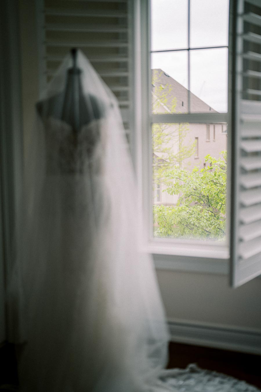 Elegant white bridal gown on mannequin next to window with outdoor view of a house and green tree leaves through open shutters, creating a serene pre-wedding ambiance.