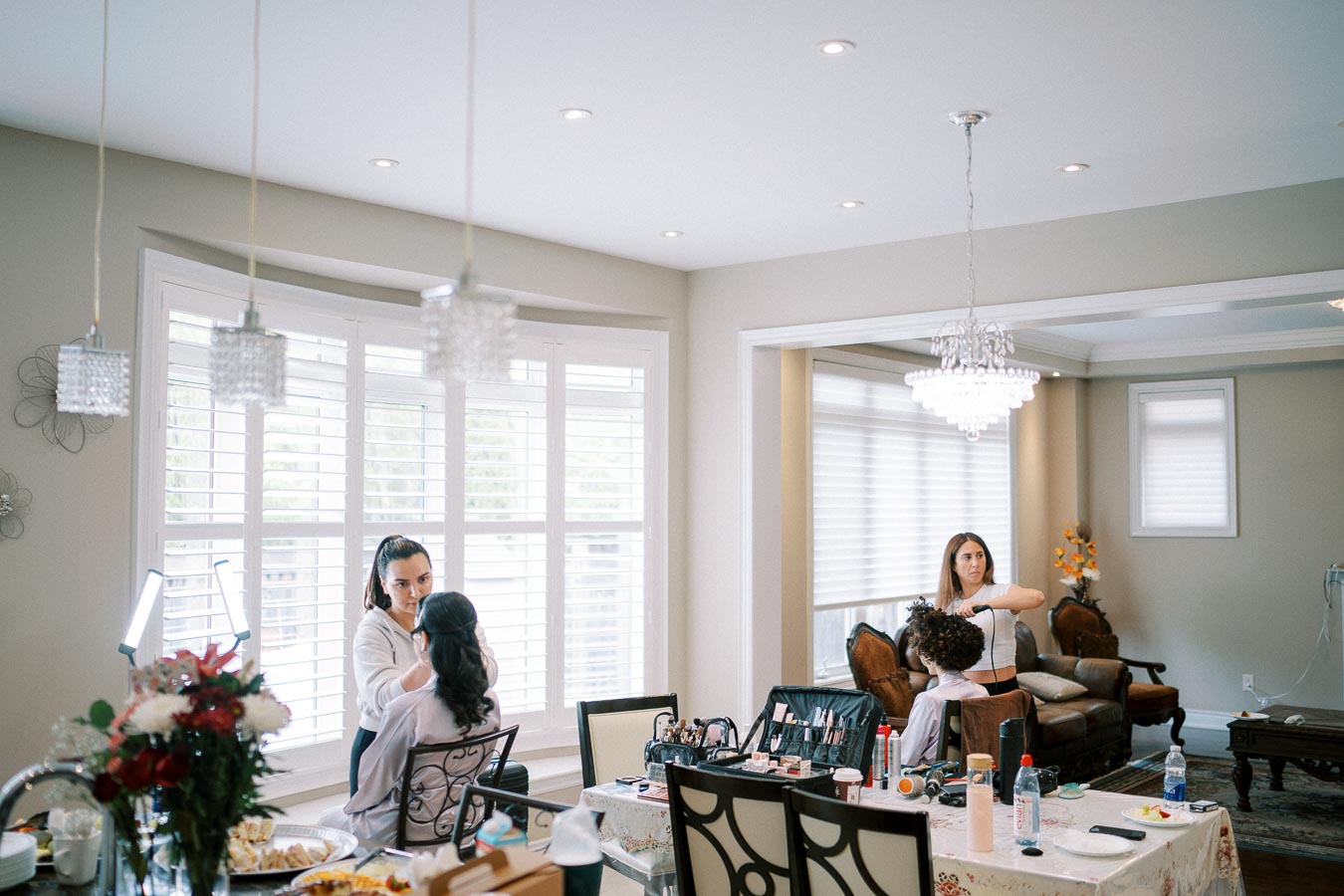 Elegant room setup with professional hair and makeup artists preparing individuals for an event. The setting features large windows, a chandelier, and organized beauty tools, creating a glamorous and sophisticated atmosphere.