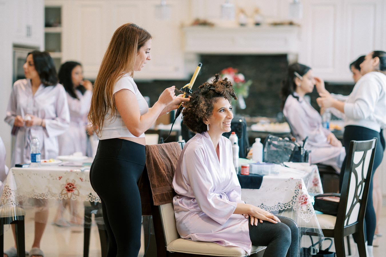 Group of women getting ready in a dressing room, with a hairstylist curling hair of a smiling woman in a robe, surrounded by beauty products and styling tools, capturing a lively and joyful atmosphere.