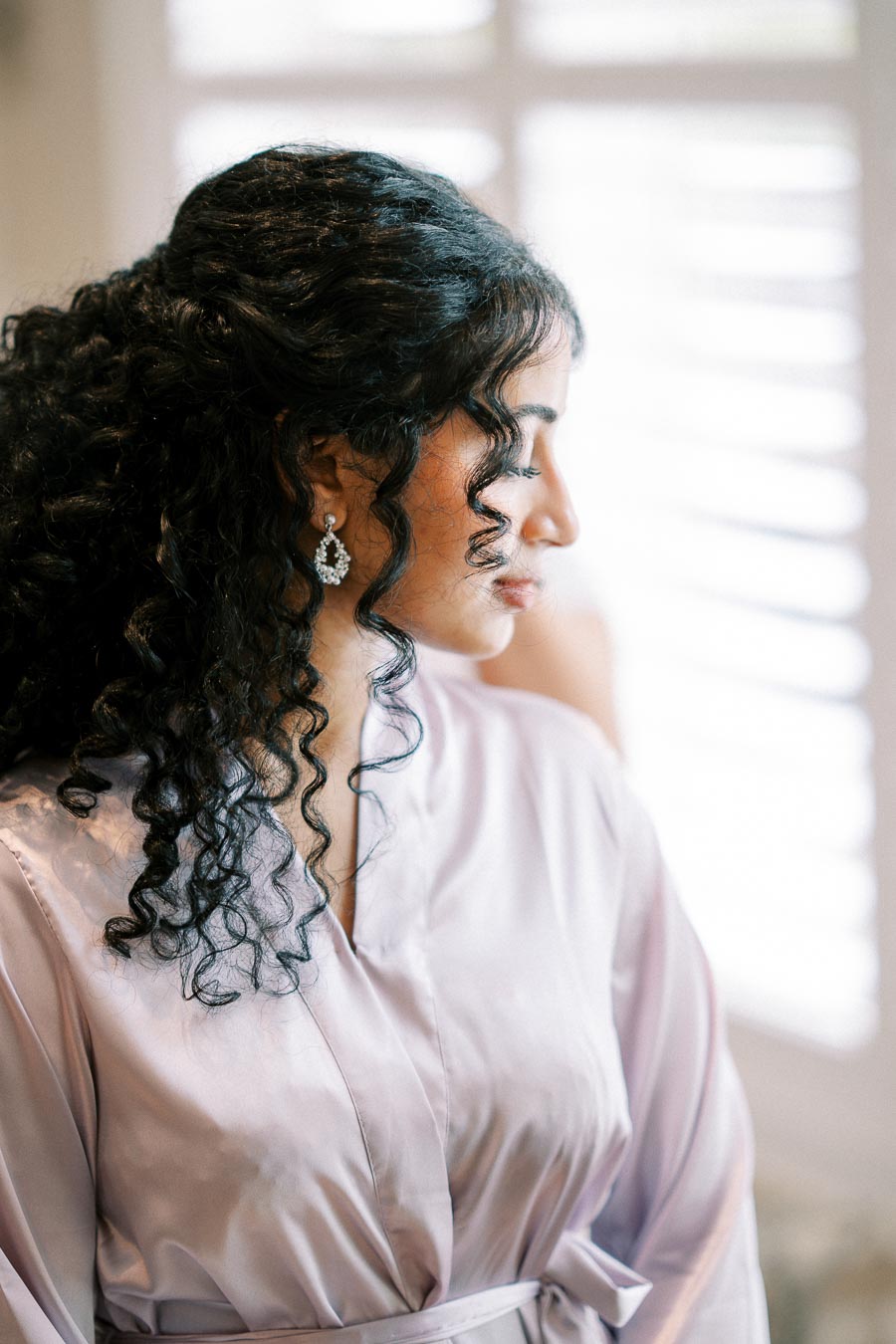 A woman with curly hair in a soft purple satin robe, looking down, with delicate earrings, standing near a window with white blinds.