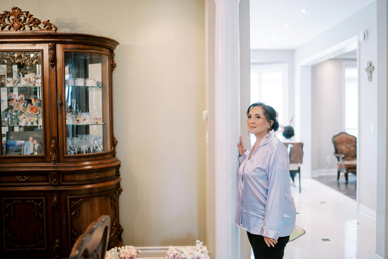 A woman in a light blue blouse stands by a white pillar in an elegantly furnished living room with a wooden display cabinet holding decorative items in the background.