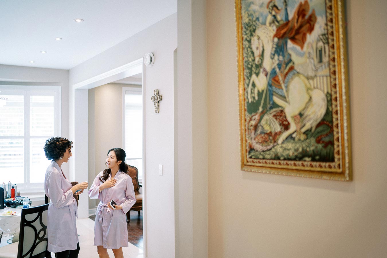 Two women in lavender robes share a conversation in a brightly lit living room, with a decorative painting and cross on the wall, creating a cozy and elegant atmosphere.