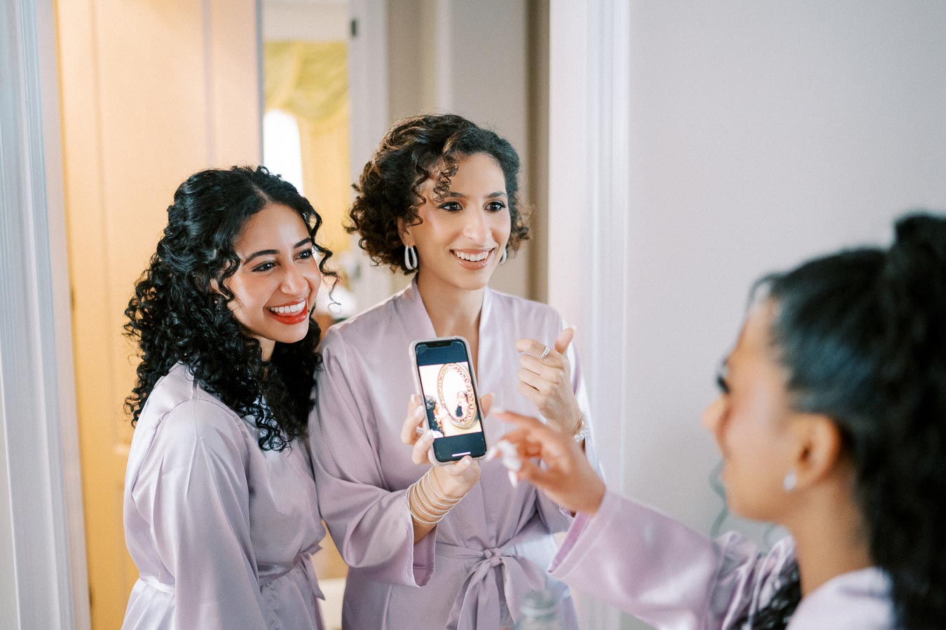 Three women in matching lavender robes share a joyful moment, capturing a selfie on a smartphone in a bright and inviting room.