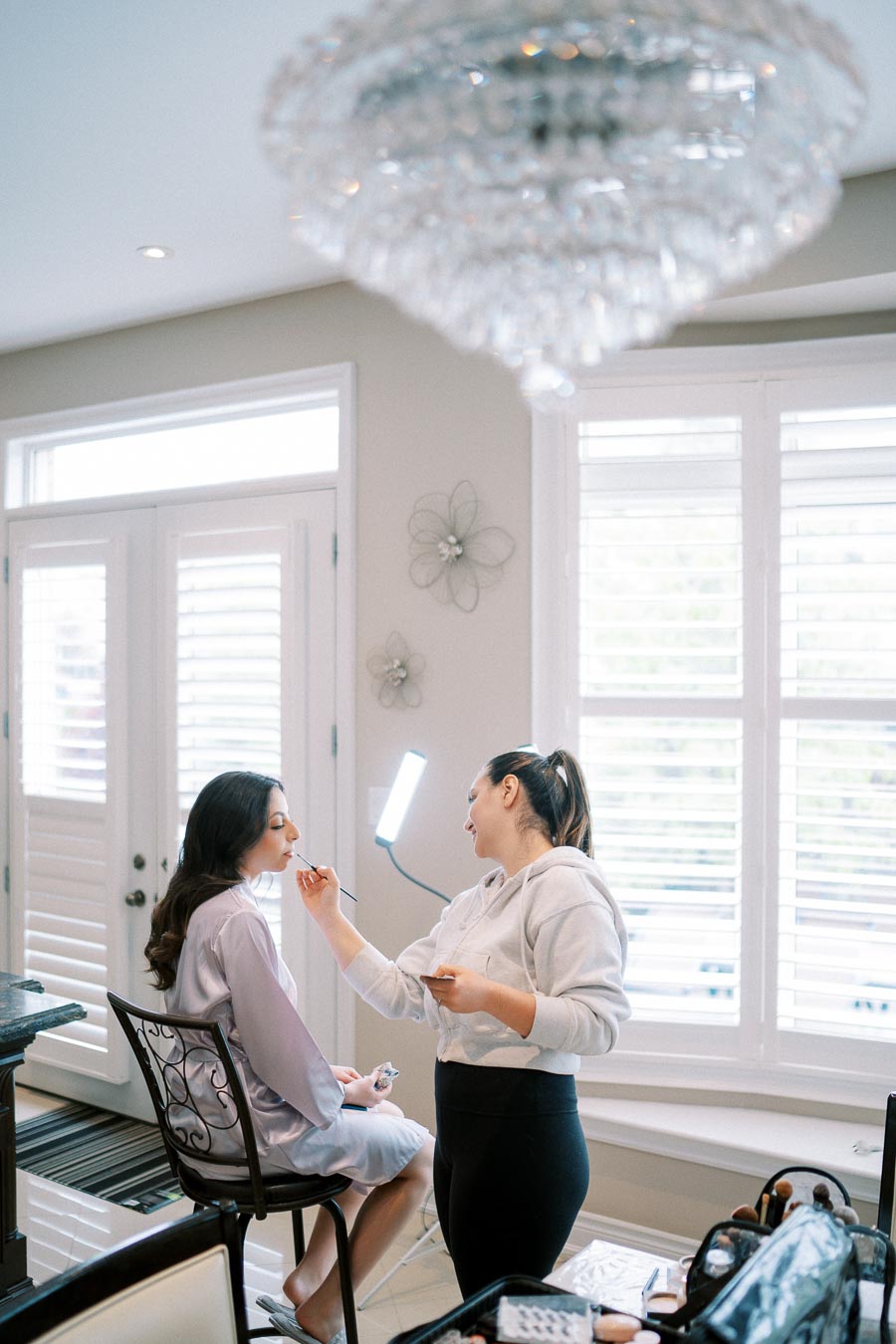 A makeup artist applies cosmetics to a woman sitting on a chair by a window in a bright room.