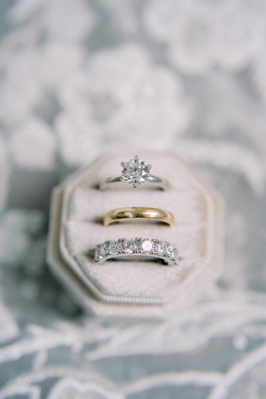 Alt text: Elegant collection of wedding rings displayed in a soft velvet box, featuring a diamond engagement ring, a gold wedding band, and a diamond eternity band arranged on a delicate lace background.