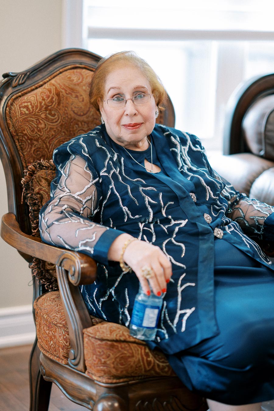 Elderly woman sitting on a patterned armchair, wearing a blue embroidered outfit and holding a water bottle, in a warmly lit room with a window in the background.