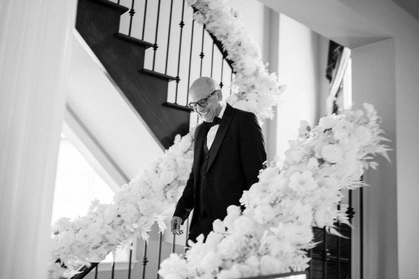 A man in a formal suit walks down a staircase elegantly decorated with white flowers, creating a serene and celebratory atmosphere.