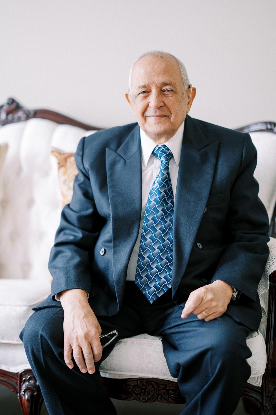 Elderly man in a dark suit and patterned blue tie sitting on a white vintage sofa, exuding a warm smile and relaxed demeanor.