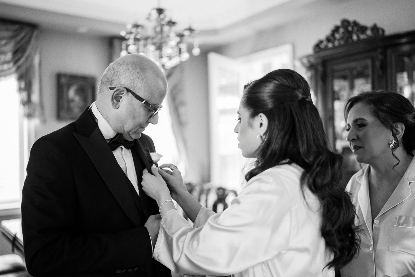 A bride adjusts a boutonniere on an older man's tuxedo while a woman looks on in a warmly lit room, possibly during a wedding preparation.