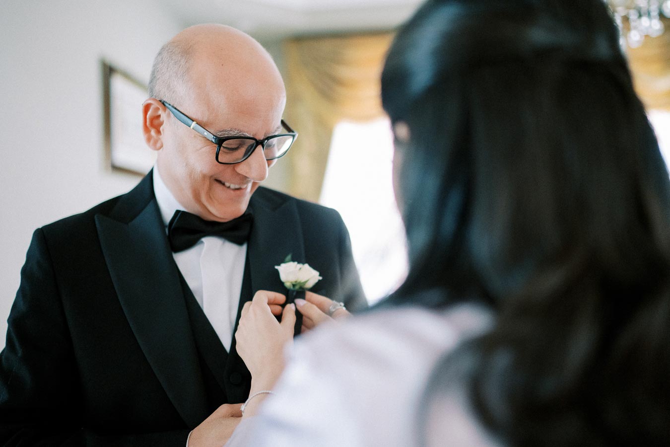 A man in a tuxedo smiling as a woman adjusts his boutonniere, preparing for a formal event in a warmly lit room.