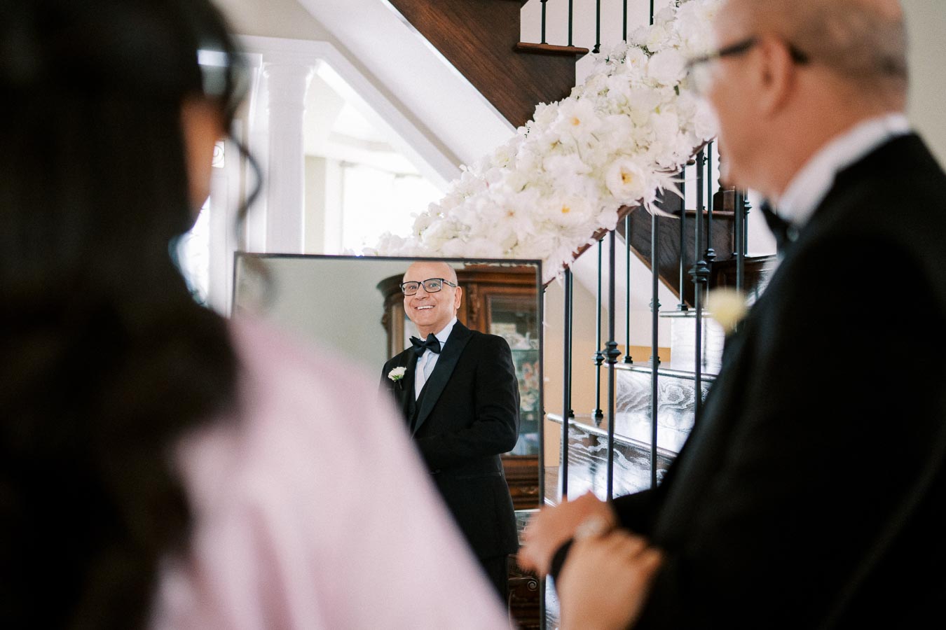 A man in a tuxedo smiling at his reflection in a mirror, with flower decorations on the staircase in the background, as a woman looks on, dressed for a formal event.