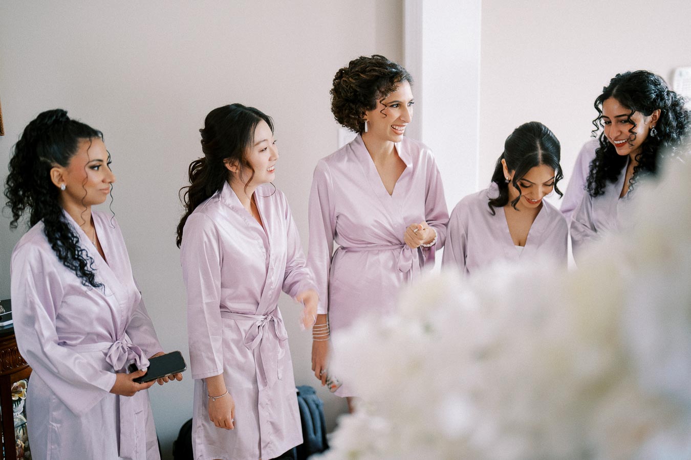 Five women in matching light purple robes smile and laugh together indoors, possibly during a bridal preparation or girls' event, creating a joyful and intimate atmosphere.