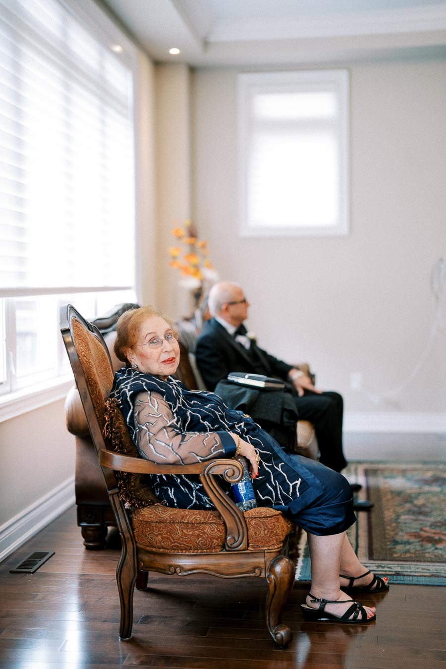 Elderly woman in elegant attire sitting on an ornate chair in a well-lit room, with a man in a suit sitting behind her.