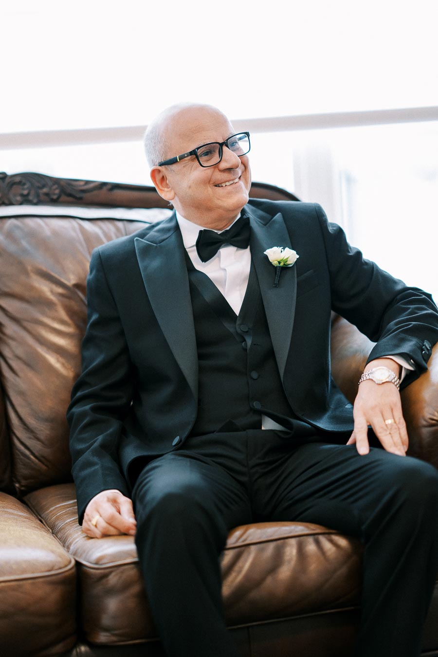 A smiling man in a tuxedo sitting on a brown leather sofa, wearing glasses and a boutonniere, exuding elegance and sophistication.