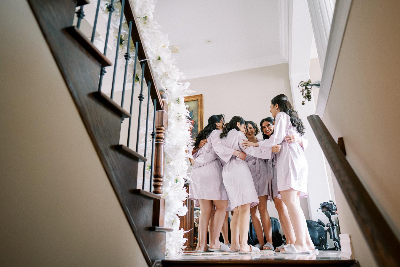 Bridesmaids in lavender robes share a group hug at the top of a decorated staircase on the morning of a wedding celebration.
