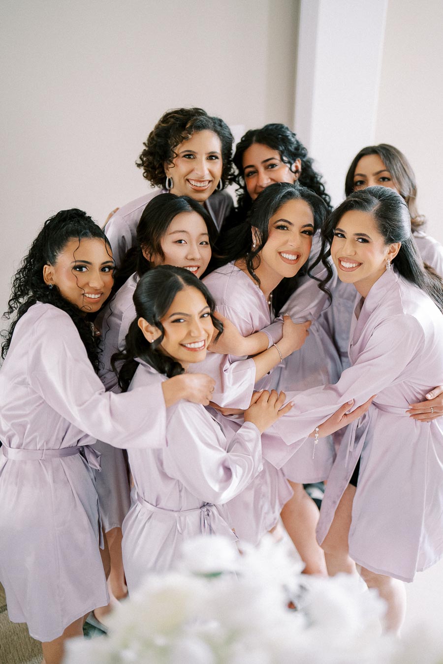 Group of bridesmaids smiling and embracing, wearing matching lavender robes on a wedding day.
