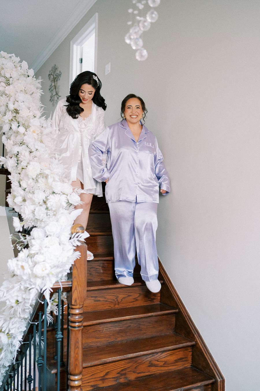 Two women in elegant satin robes descend a wooden staircase decorated with white flowers, creating a joyful and serene atmosphere.