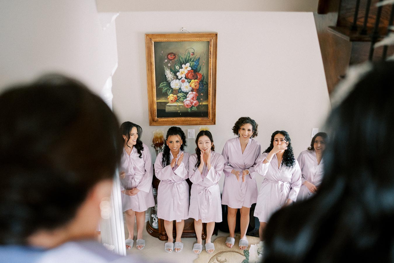 A group of six women in matching lavender robes stands smiling and excited in a room beside a large painting, preparing for a special event, viewed from behind another person.