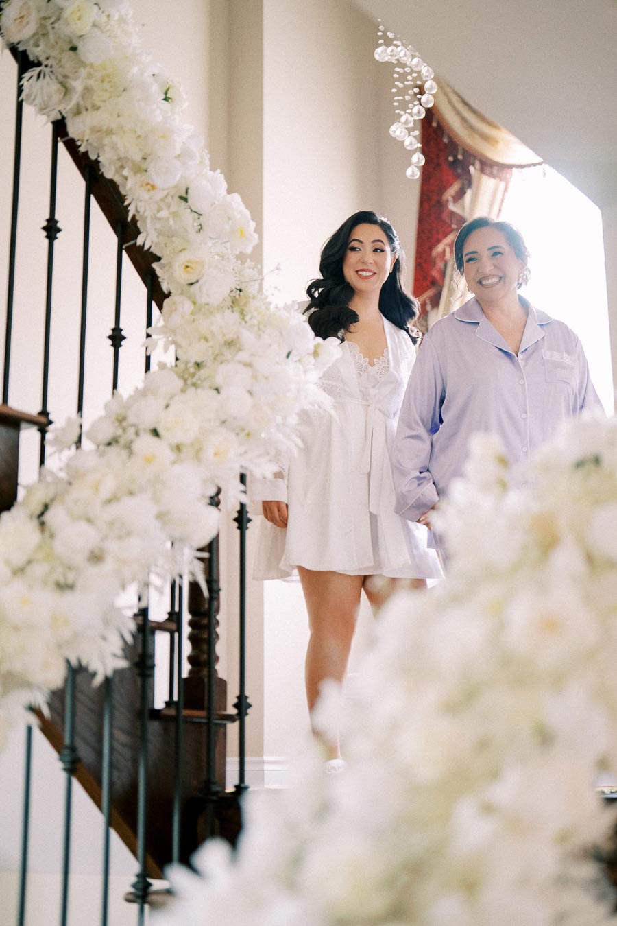 Two women smiling as they descend a staircase adorned with white floral decorations, with soft natural light illuminating the scene, creating an elegant and joyful atmosphere.