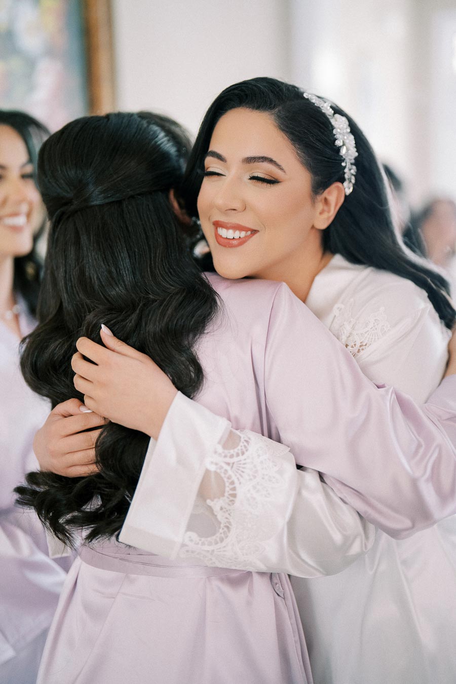 Bride embracing bridesmaid with a joyful smile, wearing decorative hairpiece and elegant satin robes, capturing a heartfelt moment of friendship and celebration.