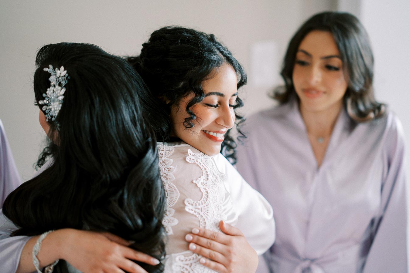 Three women wearing elegant satin robes, one smiling as she embraces another, showcasing a joyful and intimate moment likely during a wedding preparation.