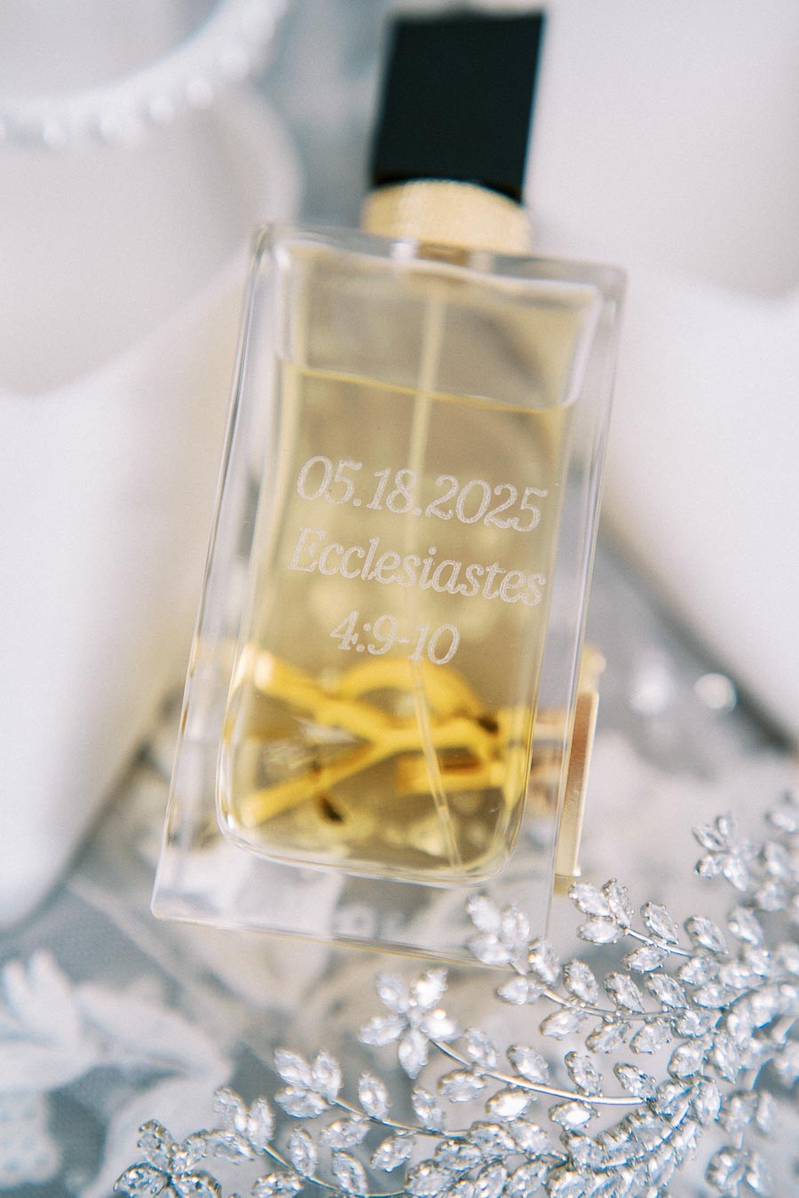 A close-up of a perfume bottle with a black cap on a decorative floral surface. The bottle is engraved with the date 05.18.2025 and a Bible verse reference Ecclesiastes 4:9-10, designed as an elegant wedding keepsake.