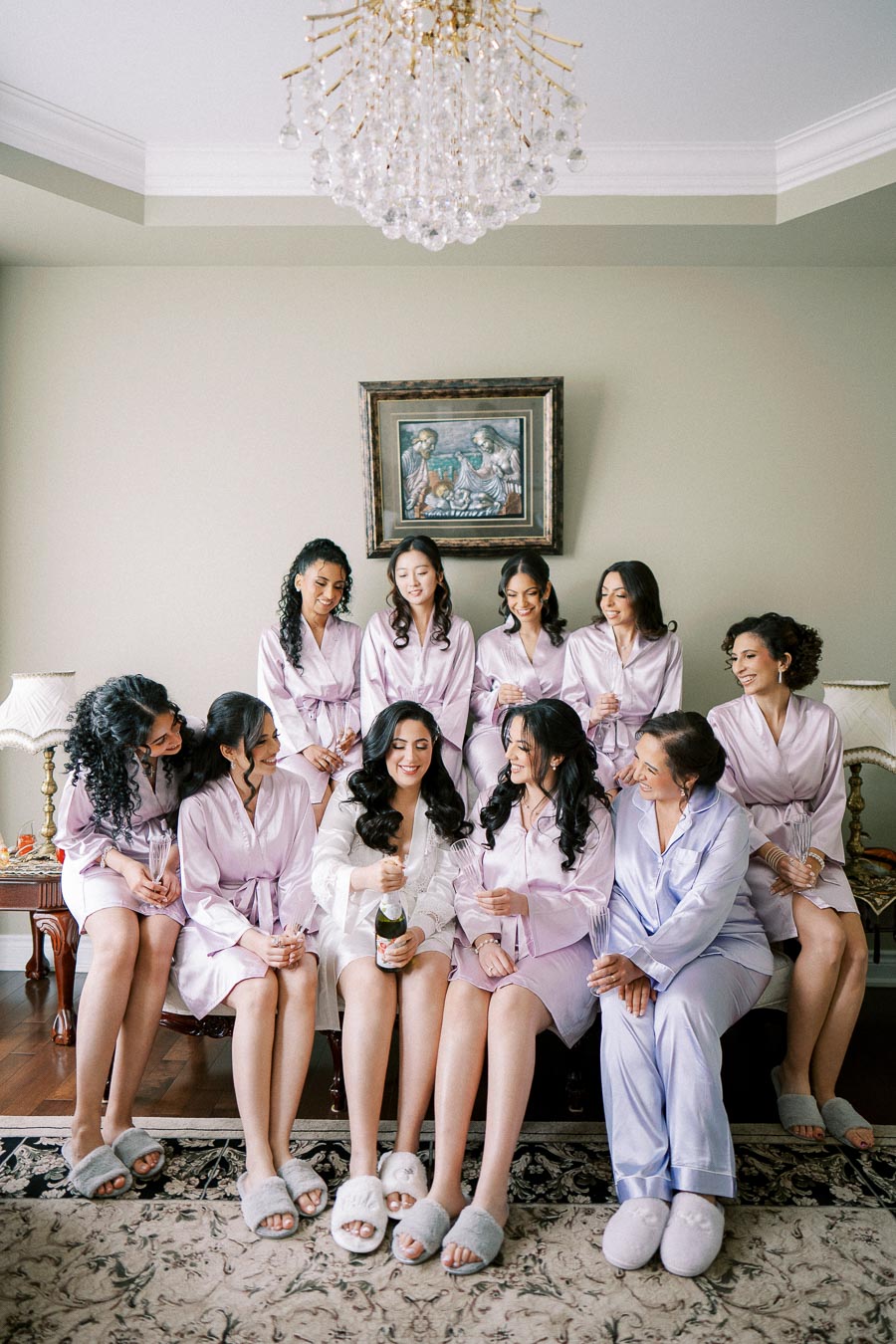 A group of women in matching lavender and white robes, smiling and sitting together on a couch, with one holding a champagne bottle in a cozy room with elegant decor, preparing for a wedding.