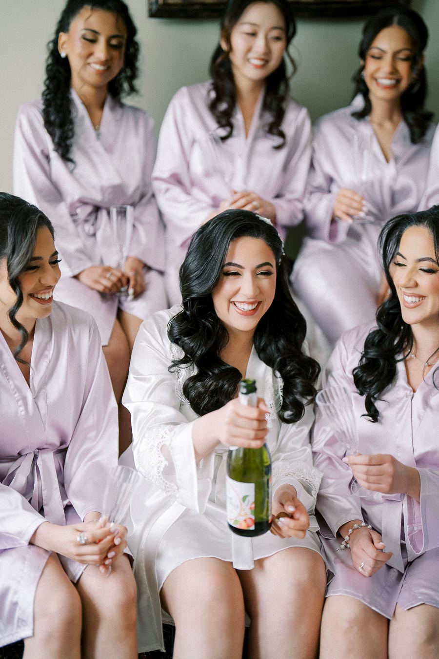 Bridal party in matching satin robes joyfully preparing for a wedding, with the bride holding a champagne bottle surrounded by her bridesmaids.