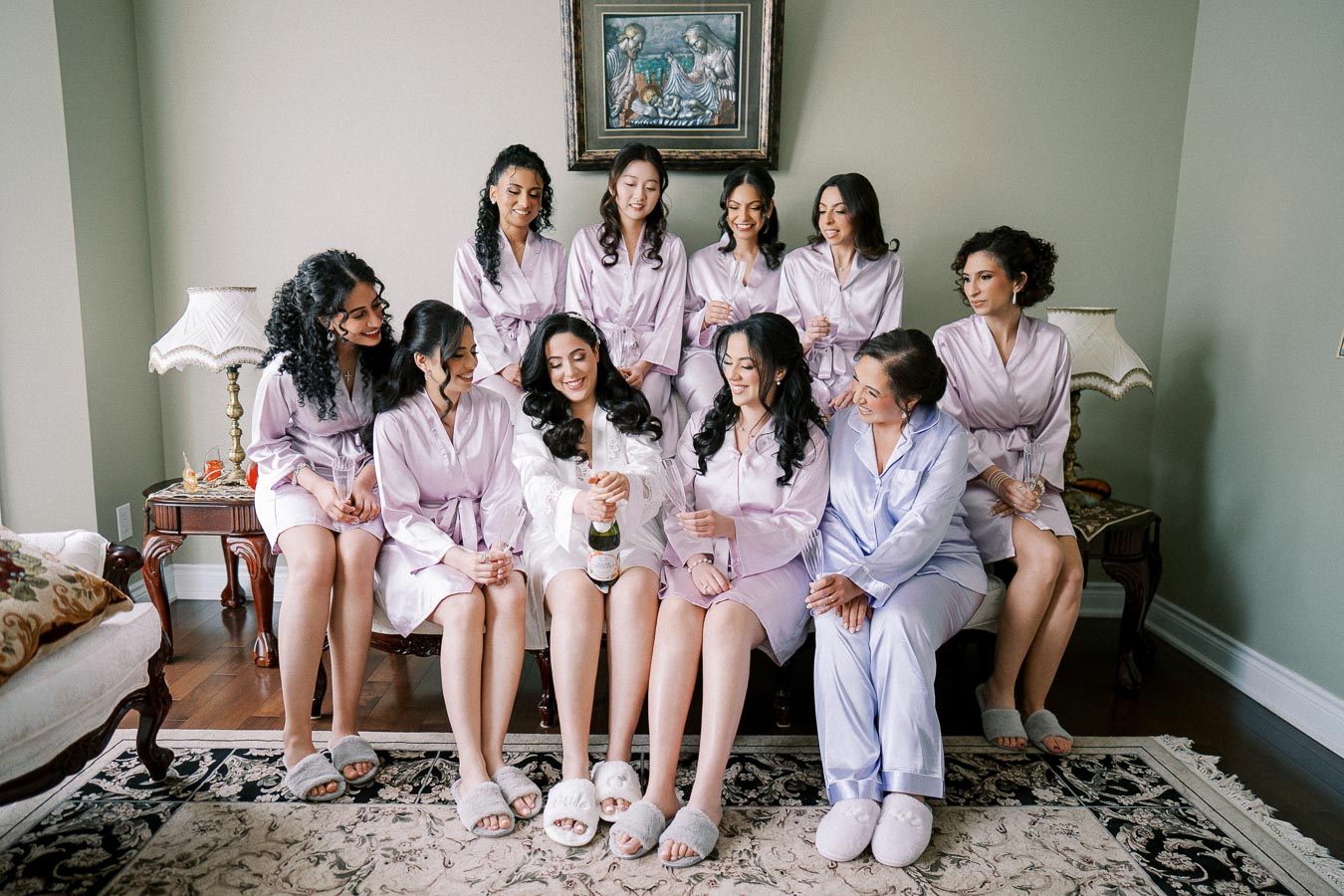 Group of women in matching robes sitting together, sharing a joyous moment with champagne on a cozy rug in a warmly lit room.
