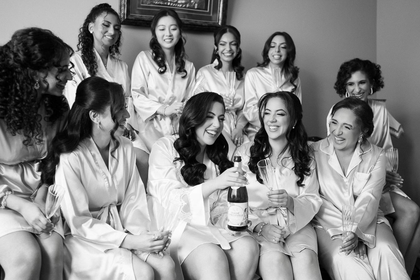 A group of women wearing matching satin robes share a joyful moment while celebrating with champagne, showcasing a festive pre-wedding atmosphere.