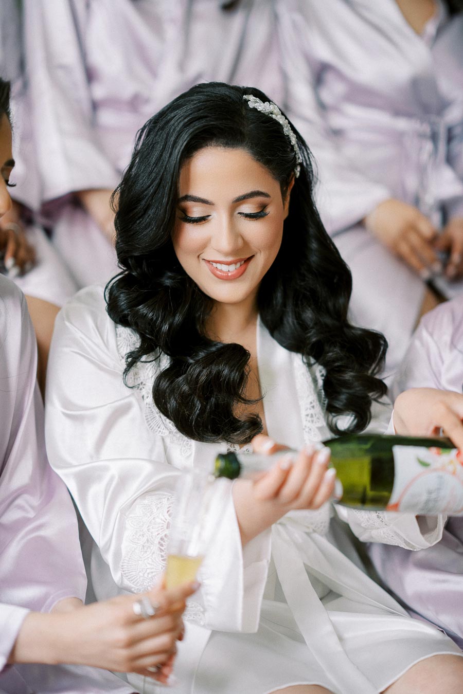 Bridal party celebrating with champagne, woman with long dark hair in white robe pouring a drink, surrounded by bridesmaids in lavender robes, joyful and festive atmosphere.