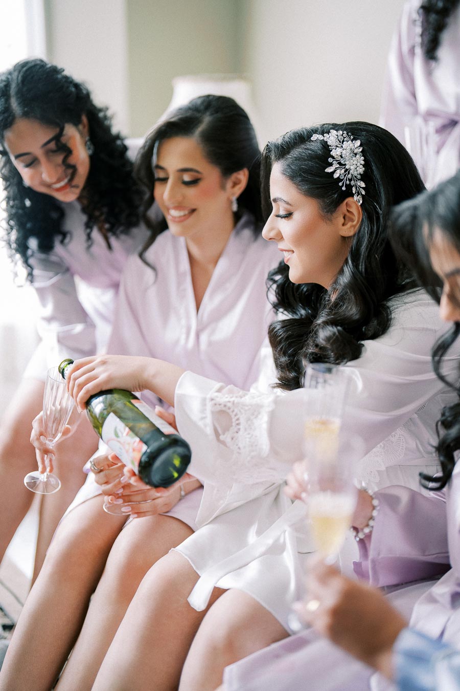 Bridal party enjoying a celebratory moment as the bride pours champagne for her bridesmaids, all wearing matching robes and smiling, creating a joyful and elegant pre-wedding atmosphere.