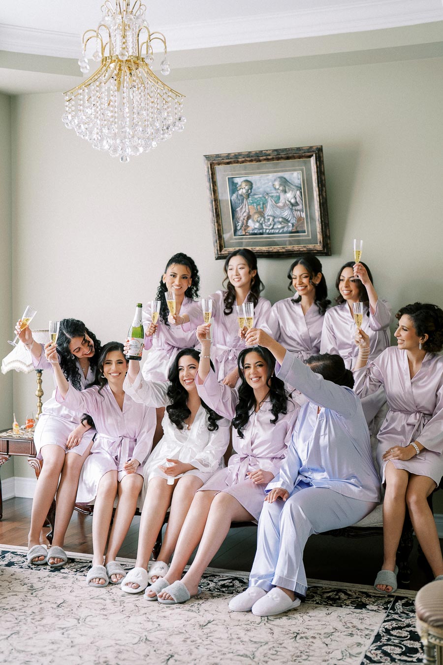 A group of women in matching light purple robes and slippers sitting in a living room, toasting with champagne glasses, under an elegant chandelier.