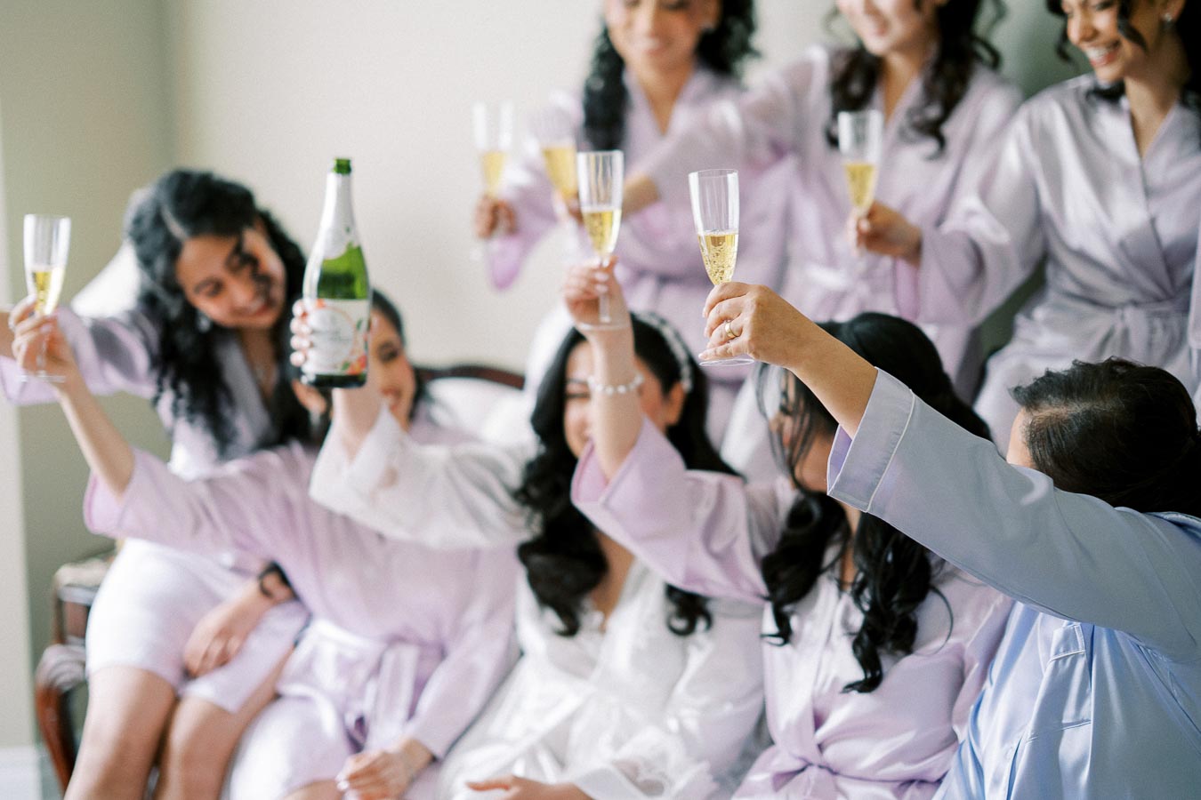 Group of women in matching lavender robes celebrating with champagne, toasting in a joyful pre-wedding preparation setting.