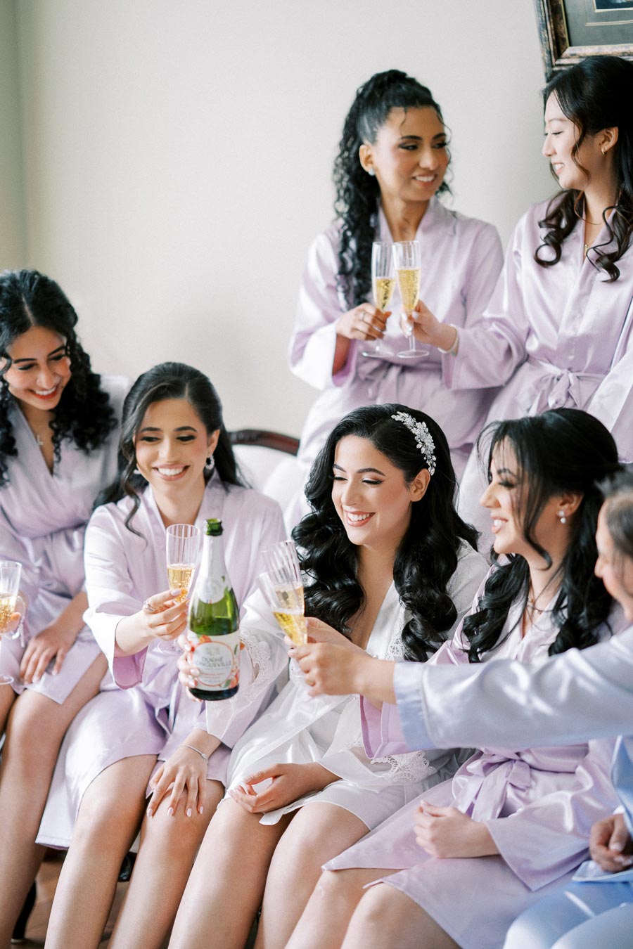Bridesmaids in matching light purple robes joyfully toasting with champagne flutes during a pre-wedding celebration.
