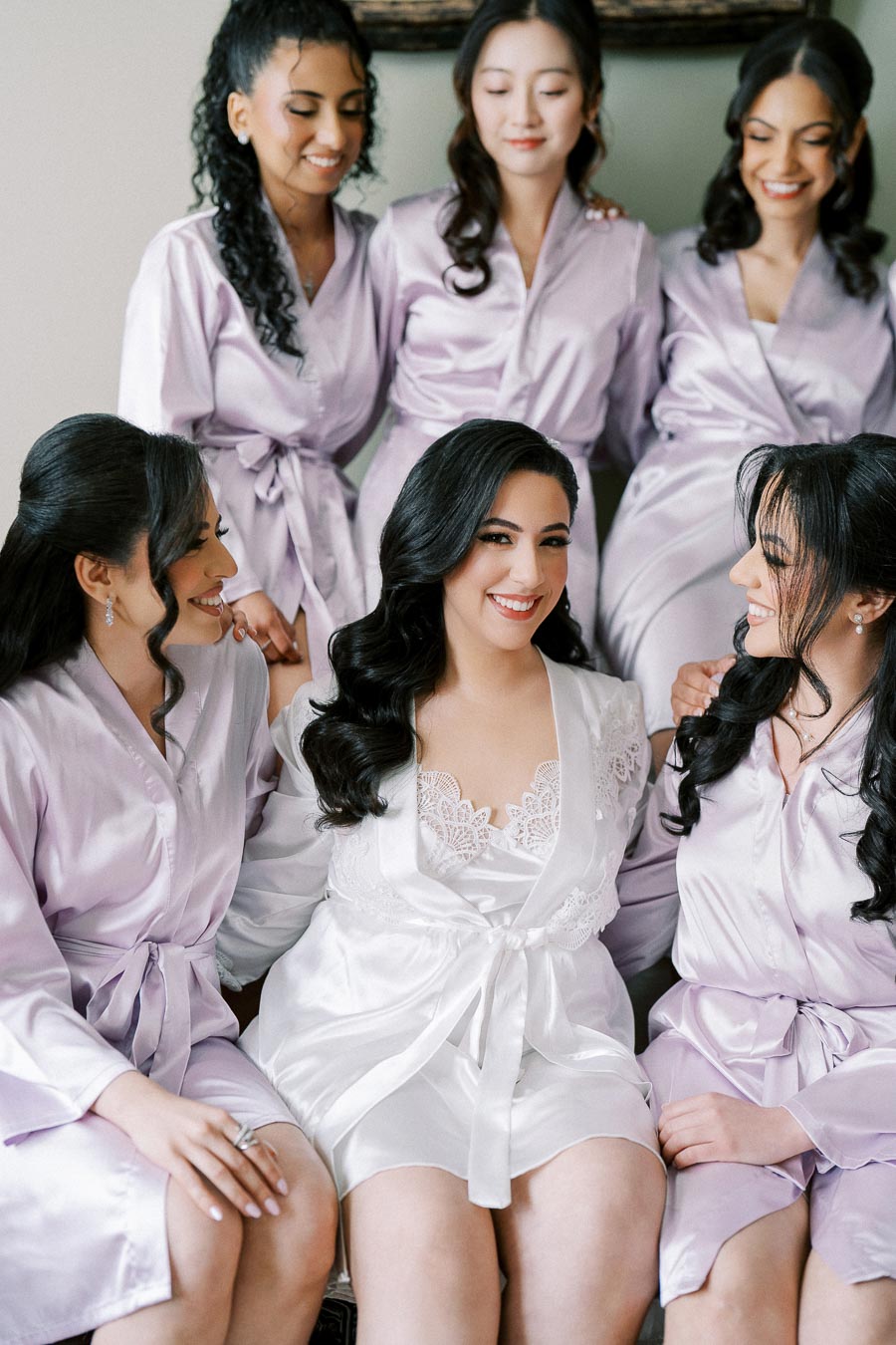 A bride in a white robe sits with her bridesmaids dressed in matching lilac satin robes, all smiling and sharing a joyful moment together before the wedding.