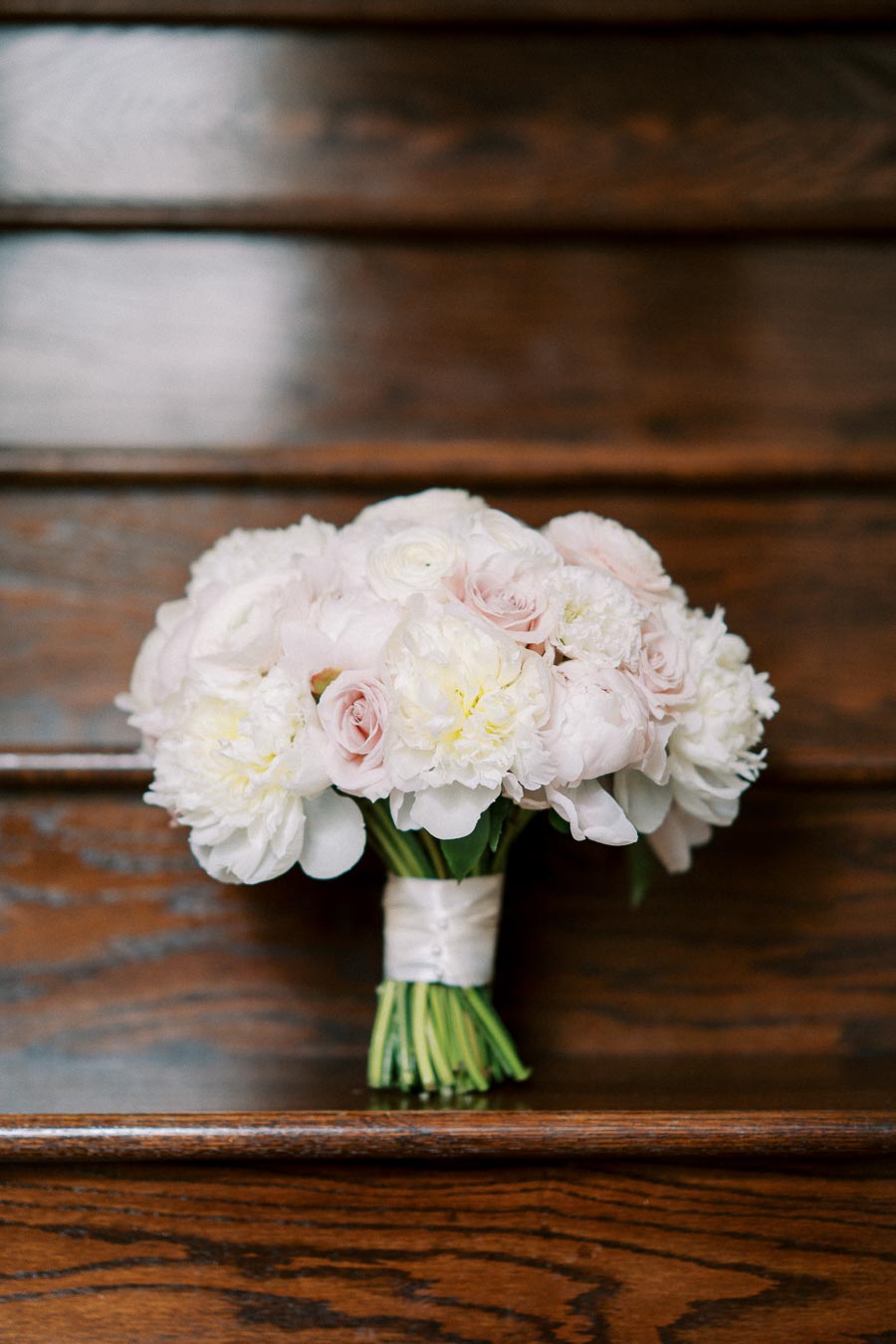 Bridal bouquet with white and pale pink roses and peonies, elegantly arranged and placed on wooden stairs.