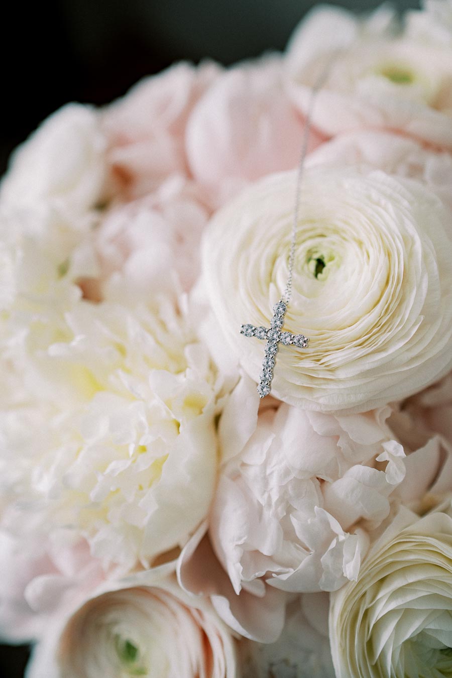 Delicate diamond cross necklace resting on a soft bouquet of white peonies and ranunculus flowers, symbolizing elegance and spirituality.