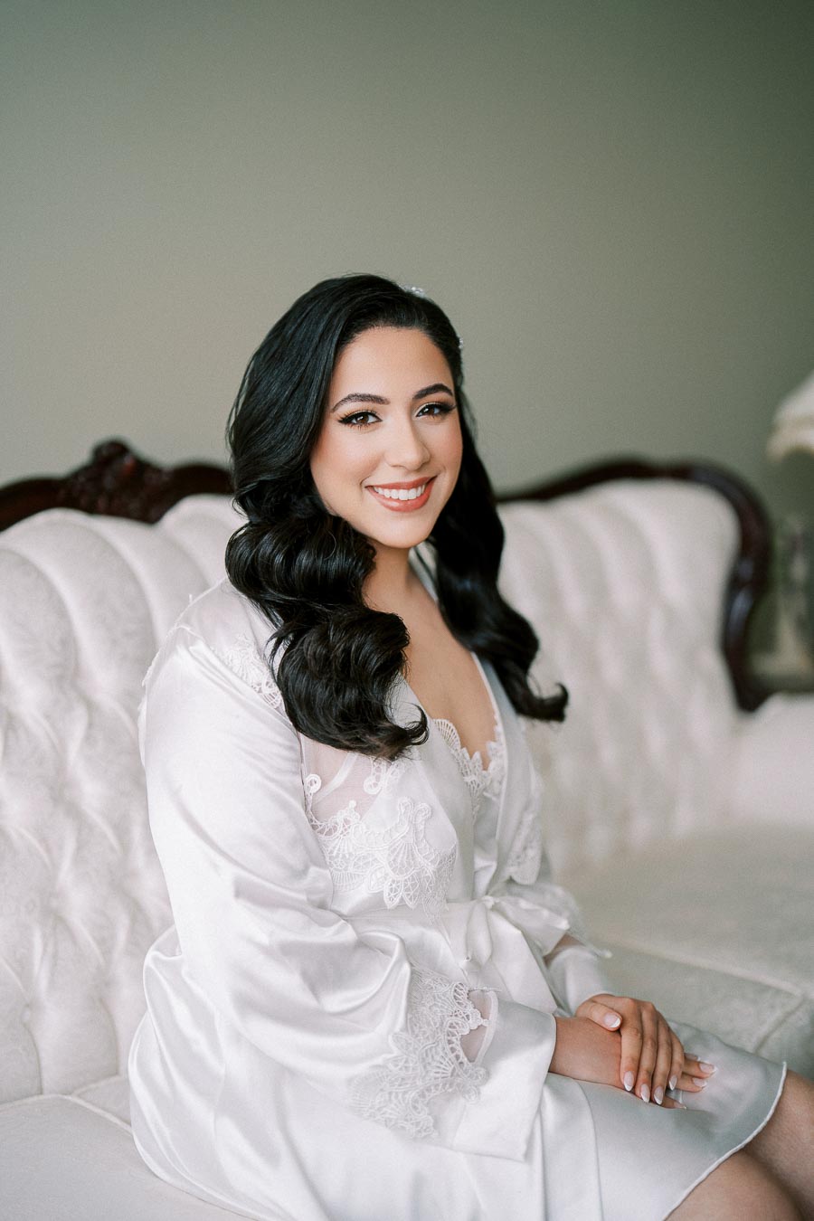 Young woman sitting on an elegant white tufted sofa, wearing a white silk robe with lace details, smiling with wavy dark hair.